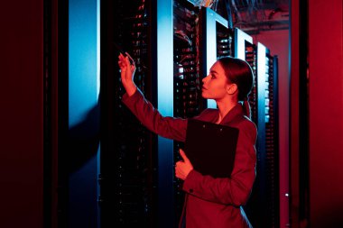 side view of businesswoman holding clipboard and looking at server room  clipart