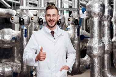 cheerful engineer in glasses and white coat standing with hand in pocket and showing thumb up  clipart
