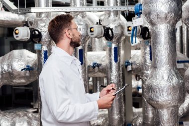 bearded engineer in white coat holding pen and clipboard near air compressed system  clipart