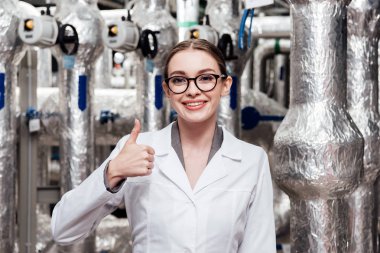 happy engineer in white coat and glasses showing thumb up near air compressed system  clipart