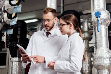 handsome engineer pointing with finger at digital tablet near attractive coworker and air compressor system  clipart