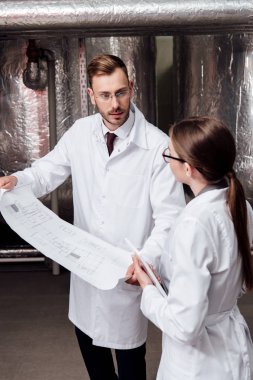 engineer in white coat looking at coworker while holding blueprint  clipart