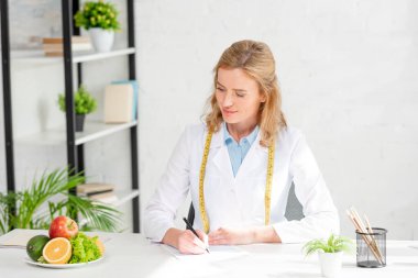 attractive nutritionist sitting at table and looking at fruits in clinic  clipart