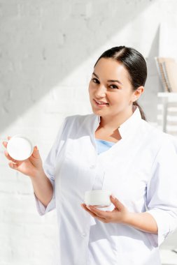 Gülümseyen dermatolog kozmetik kremi tutuyor ve klinikteki kameraya bakıyor. 