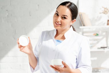 Gülümseyen dermatolog kozmetik kremi tutuyor ve klinikteki kameraya bakıyor. 