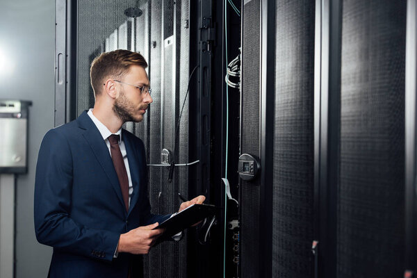 selective focus of handsome businessman in glasses and suit holding clipboard and pen in data center 