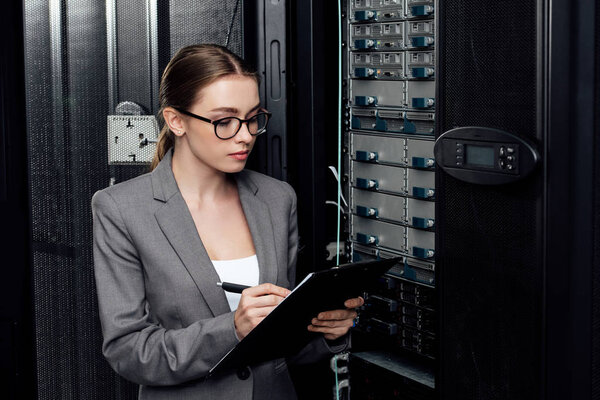 businesswoman in glasses holding clipboard while writing in data center 