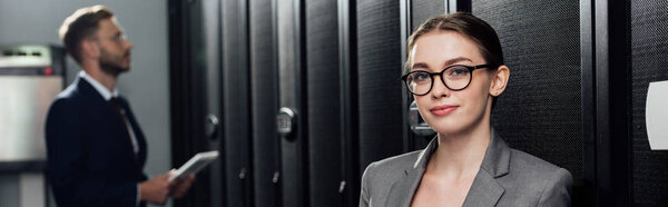 panoramic shot of attractive businesswoman in glasses looking at camera near bearded businessman in data canter 