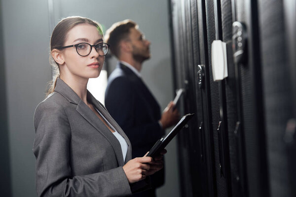 selective focus of attractive businesswoman in glasses holding clipboard near bearded businessman in data canter 