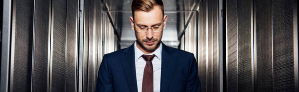 panoramic shot of businessman in glasses near server racks 