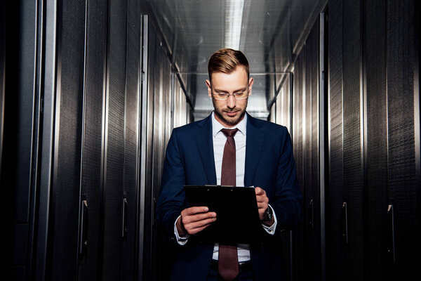 handsome businessman in glasses looking at clipboard near server racks 