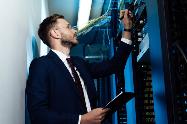 side view of bearded businessman in glasses holding pen and clipboard while looking at server rack 