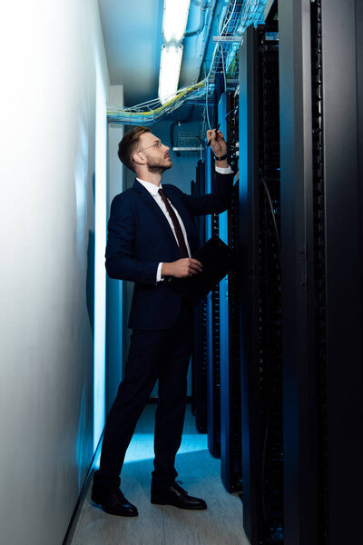 вид сбоку на красивого бизнесмена в очках, держащего ручку и буфер обмена, глядя на стойку сервера
 