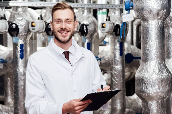 bearded and cheerful engineer in white coat holding pen and clipboard near air compressed system 