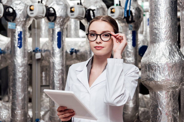 attractive engineer in white coat touching glasses while holding digital tablet near air compressed system 