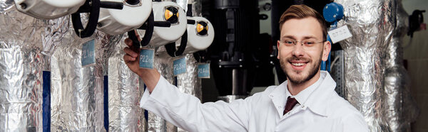 panoramic shot of cheerful bearded engineer in white coat near air supply system 