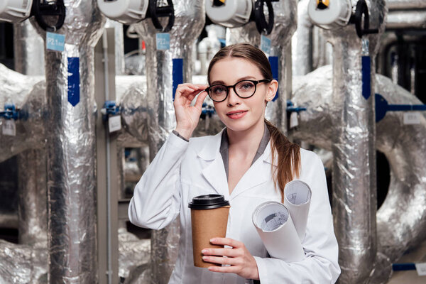 cheerful engineer in white coat and glasses holding blueprints and paper cup near air supply system 