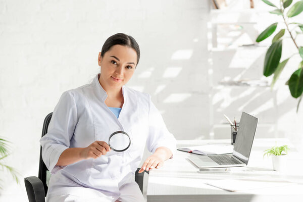 attractive dermatologist sitting at table and holding magnifying glass in clinic 