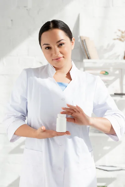 Atrakcyjny Dermatolog Białym Płaszczu Trzymający Butelkę Tabletkami Klinice — Zdjęcie stockowe
