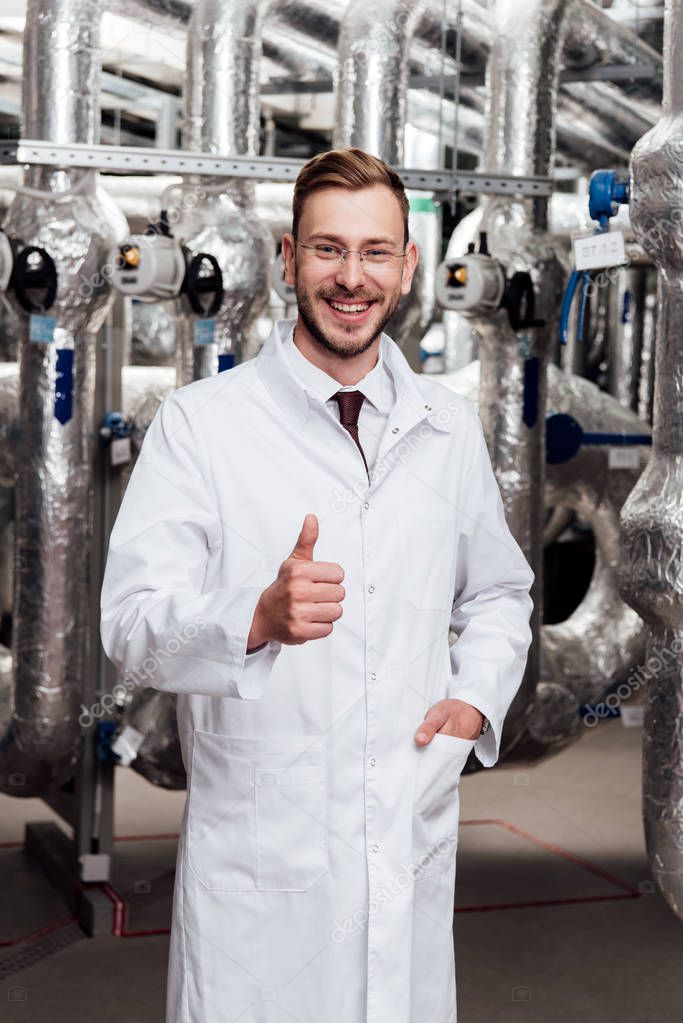 happy engineer in glasses and white coat standing with hand in pocket and showing thumb up 