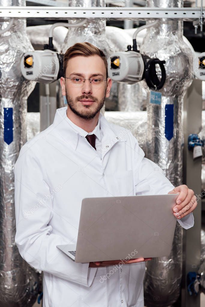 handsome bearded engineer in white coat and glasses holding laptop 