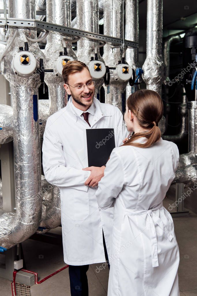 cheerful engineer looking at coworker while standing near air compressor system 