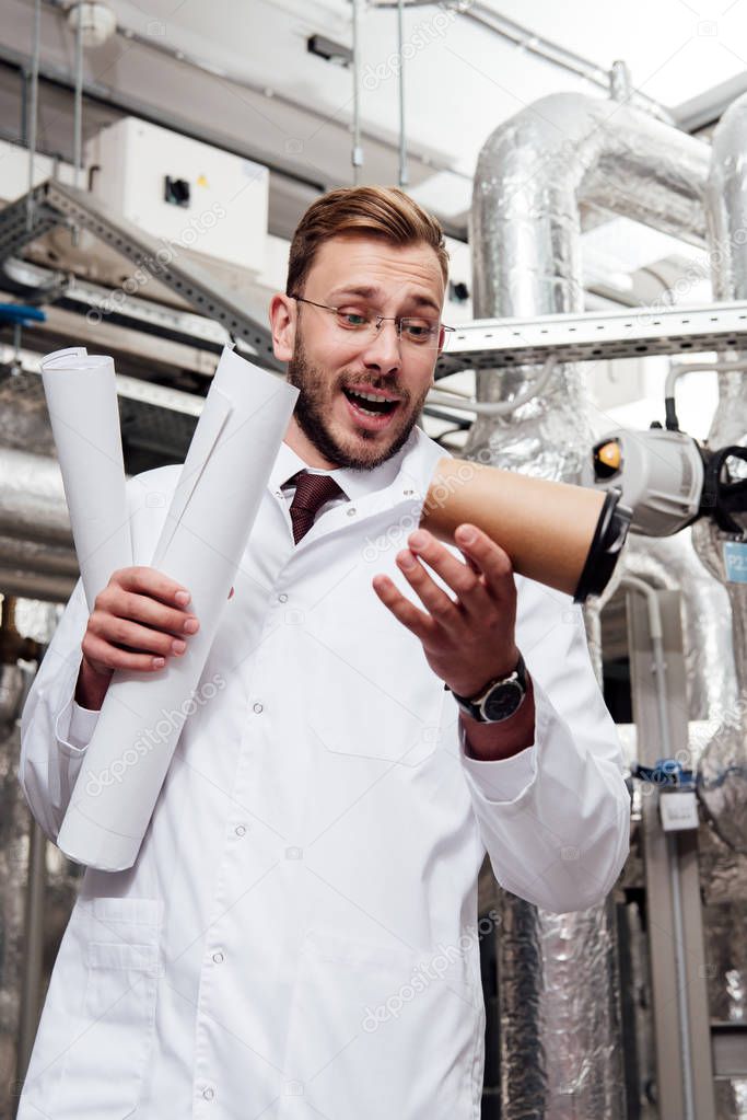worried engineer in white coat and glasses holding blueprints and looking at falling paper cup near air supply system 