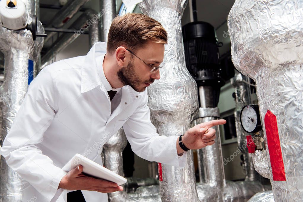 bearded engineer in white coat holding digital tablet and pointing with finger at air supply system 