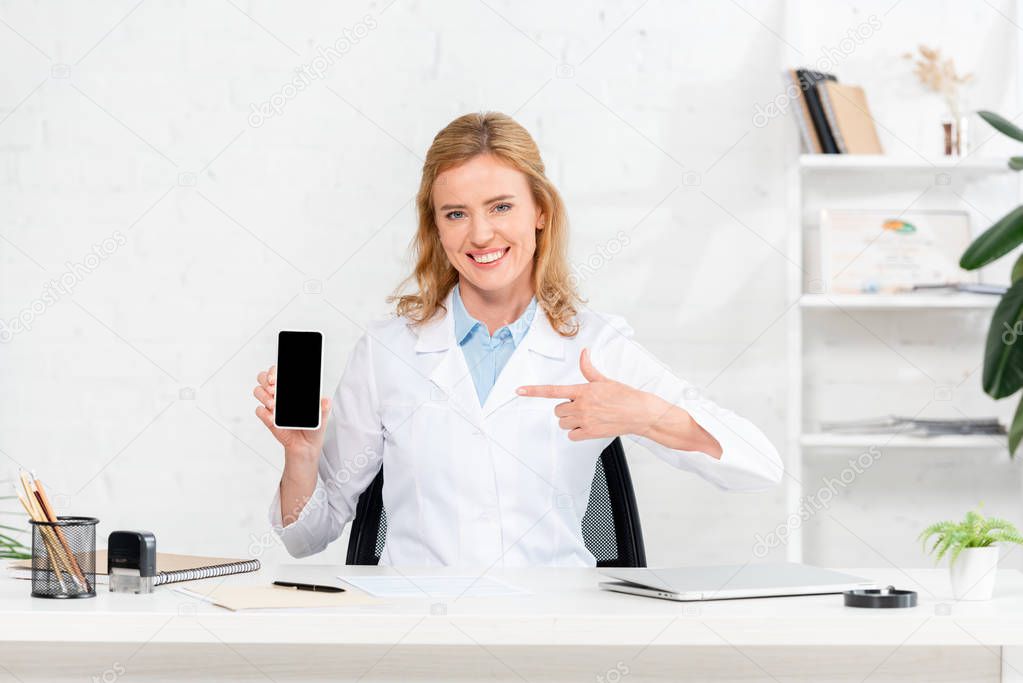smiling nutritionist sitting at table and pointing with finger at smartphone with copy space in clinic 