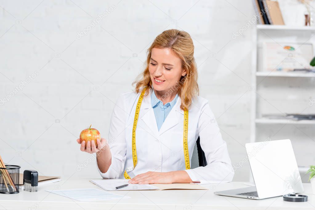 smiling nutritionist sitting at table and holding apple in clinic 