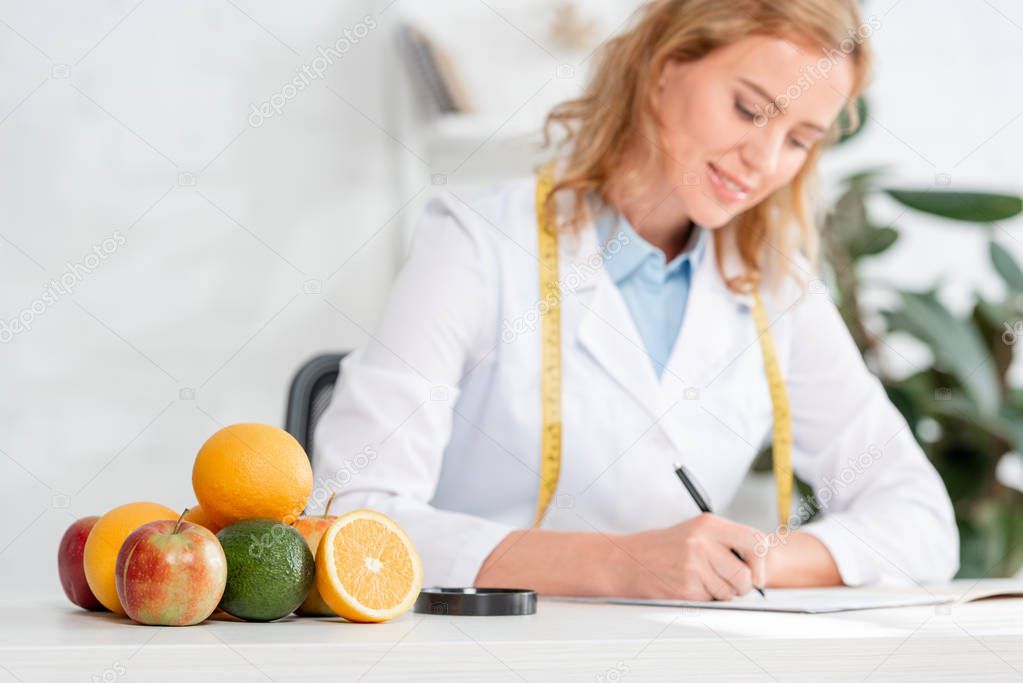selective focus of fruits and vegetable on table and nutritionist sitting at table and writing on background in clinic 