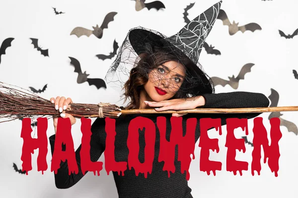 attractive woman in witch hat holding broom in Halloween near Halloween red lettering