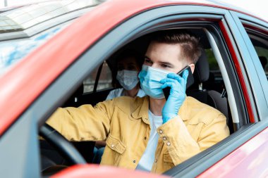 Tıbbi maskeli ve eldivenli erkek sürücü Coronavirus salgını sırasında takside yolcularla telefonda konuşuyor.