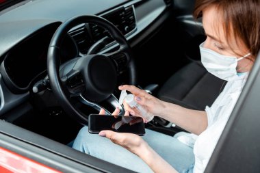 Coronavirüs salgını sırasında arabadaki akıllı telefon için antiseptik kullanan tıbbi maskeli bir kız.