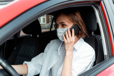 Tıbbi maskeli kadın covid-19 salgını sırasında araba kullanırken telefonla konuşuyor.