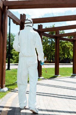 workman in hazmat suit and respirator disinfecting wooden construction in park during coronavirus pandemic clipart