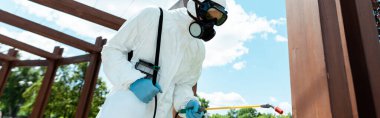 Koronavirüs salgını sırasında parktaki ahşap yapıyı dezenfekte eden tehlikeli madde giysisi ve yüz maskesi takan işçiler.