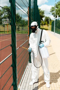 professional specialist in hazmat suit and respirator disinfecting fence of basketball court in park during coronavirus pandemic clipart