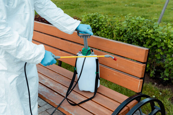 cropped view of specialist in hazmat suit and respirator disinfecting bench in park during coronavirus pandemic