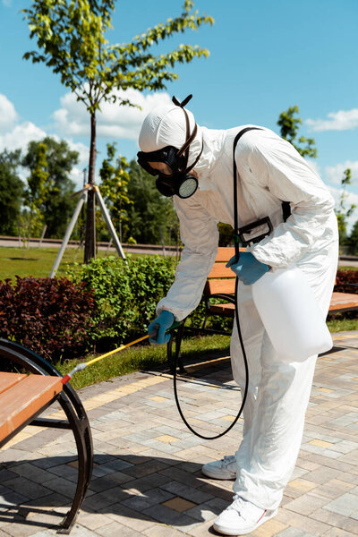 specialist in hazmat suit and respirator disinfecting bench in park during coronavirus pandemic