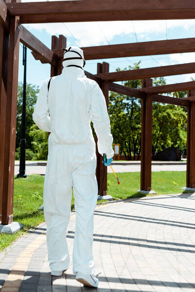 workman in hazmat suit and respirator disinfecting wooden construction in park during coronavirus pandemic