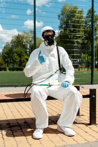 cleaning specialist in hazmat suit and respirator showing thumb up while sitting on bench with spray bag during covid-19 pandemic