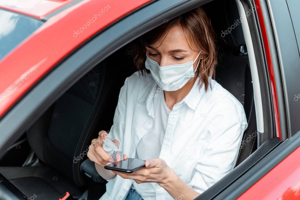 girl in medical mask using antiseptic for smartphone in car during covid-19 pandemic