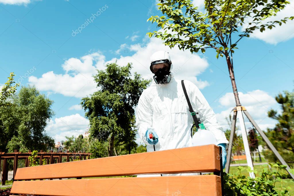 male specialist in hazmat suit and respirator disinfecting bench in park during coronavirus pandemic