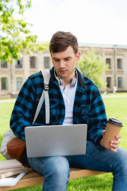 Üniversite kampüsünün yanında dizüstü bilgisayar ve kağıt bardak kullanan yakışıklı öğrenci, online çalışma konsepti 