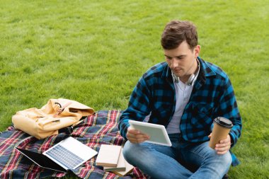 Bilgisayarın yanında dijital tablet ve kağıt bardak tutan yakışıklı öğrenci ve ekose battaniye üzerine kitaplar, online çalışma konsepti 