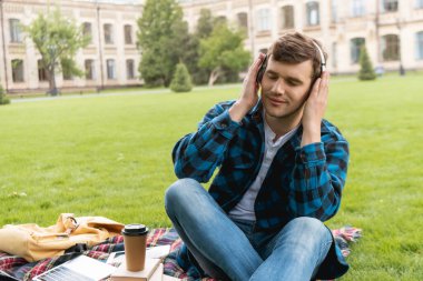 Gözü kapalı mutlu bir öğrenci kablosuz kulaklıklara dokunuyor ve laptopun yanında müzik dinliyor ve ekose battaniyede kağıt bardak kullanıyor. 