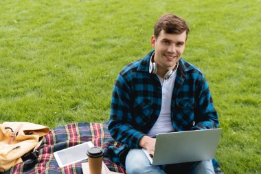 Mutlu öğrenci dijital tablet ve kağıt bardakla ekose battaniye kullanıyor, online çalışma konsepti 