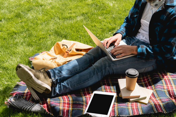 cropped view of student using laptop near digital tablet with blank screen and paper cup on plaid blanket, online study concept 