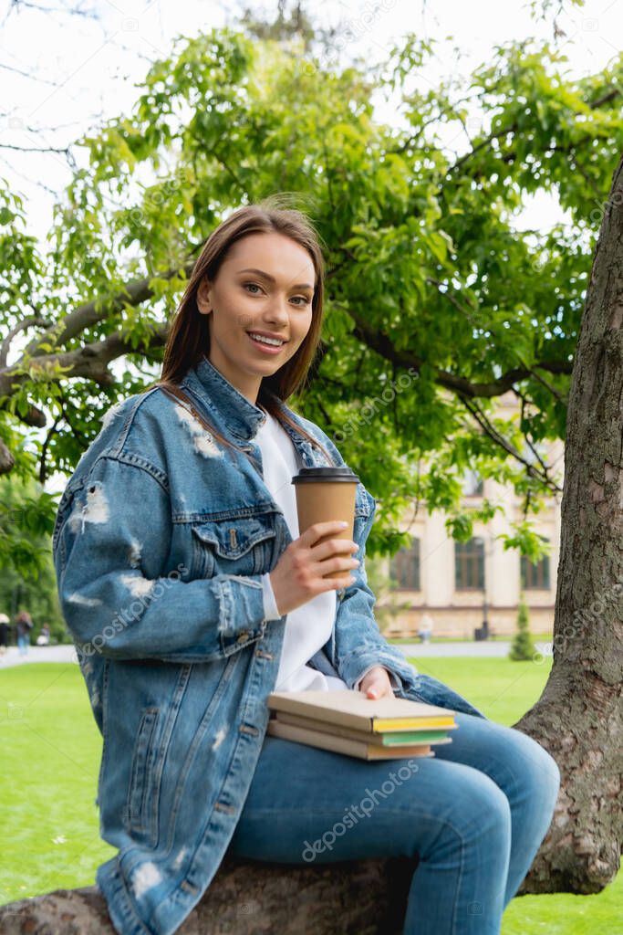 cheerful and beautiful student holding paper cup and books while sitting on tree 